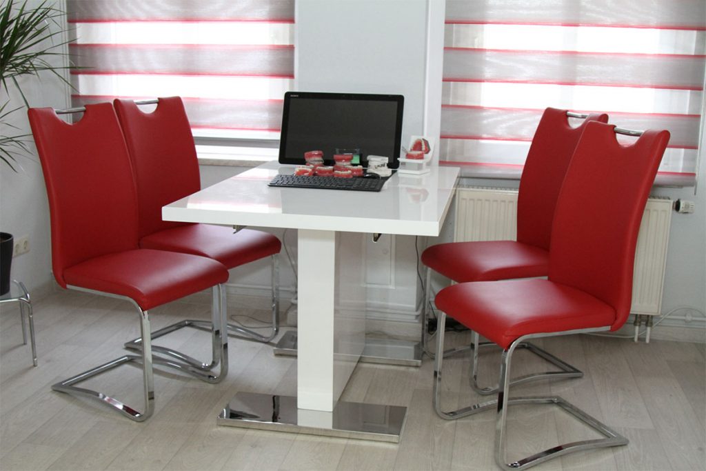 Ein Praxisbild mit einem weißen Tisch, auf dem ein Laptop steht und Beispielgebisse liegen. An ihm stehen rote Stühle mit silbernen Gestell.