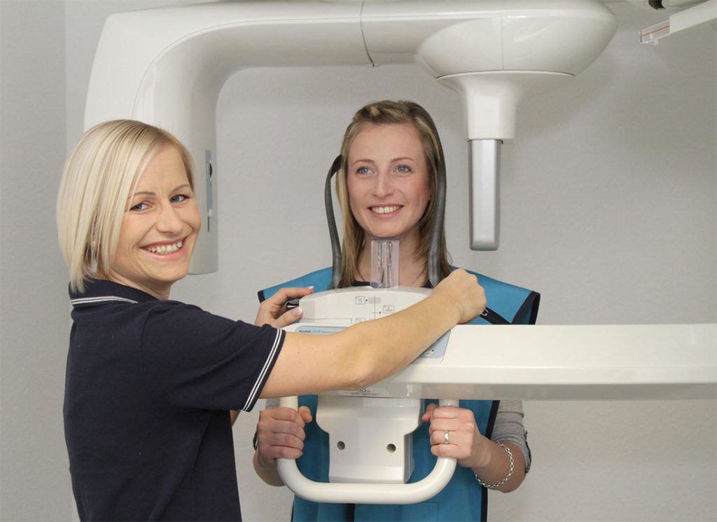 Eine Frau steht in einem Röntgengerät, eine Zahnarzthelferin blickt in die Kamera und lächelt.
