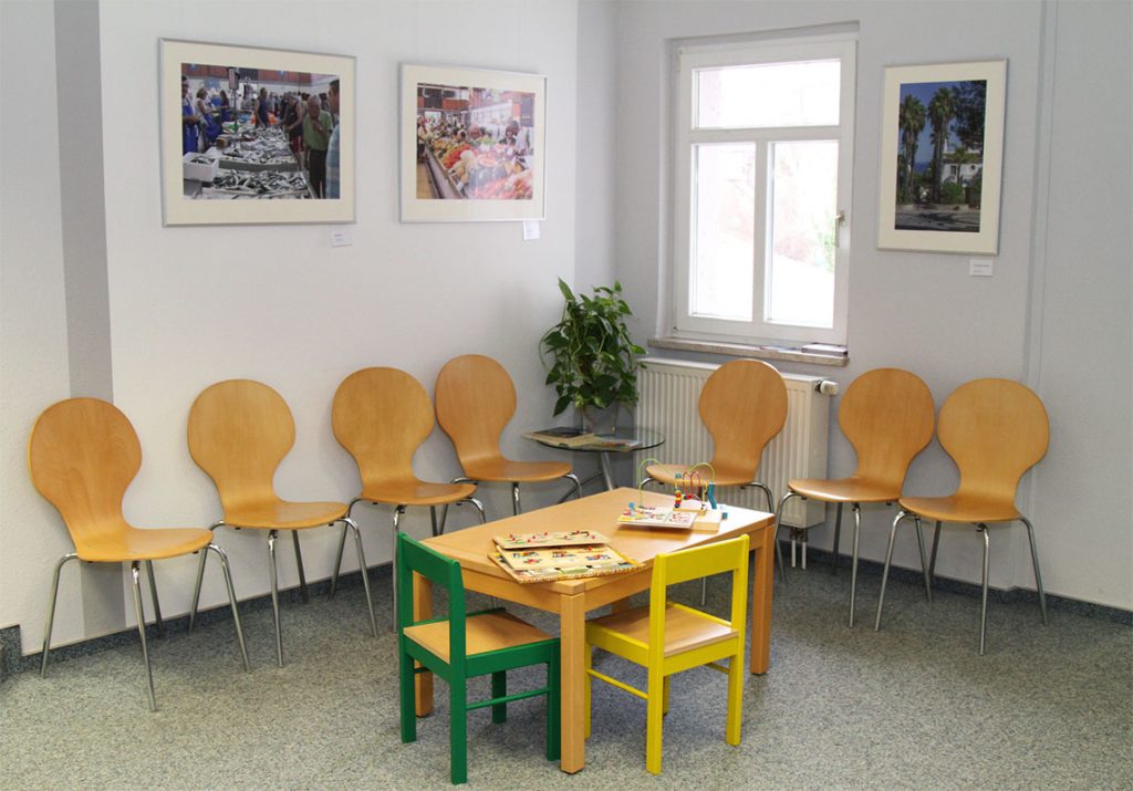 Der Warteraum mit einem Kindertisch in der Mitte, an dem ein gelber und grüner Stuhl stehen. Außen herum stehen 6 Holzstühle.